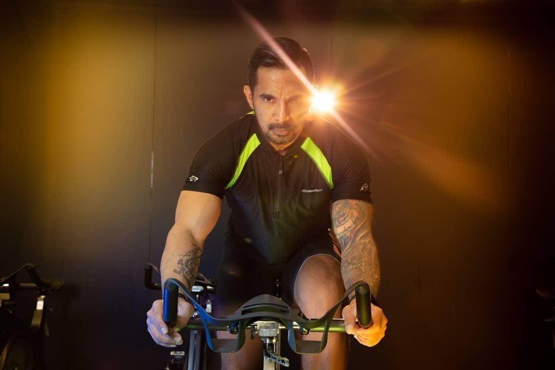 Ron Garcia riding an indoor cycle in a darkened studio, lit from behind by a single warm light
